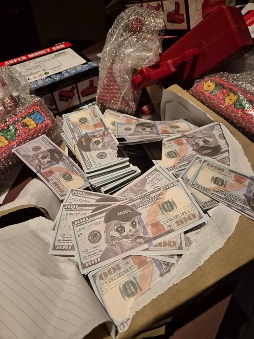 A cardboard box filled with stacks of novelty "HODL" hundred-dollar bills, surrounded by wrapped gifts including boxes labeled "SUPER MONEY GUN."