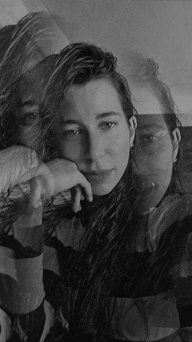 A black and white self-portrait photograph of a woman with long hair resting her chin on her hand, featuring a dreamy double-exposure effect showing multiple overlapping images of her face.