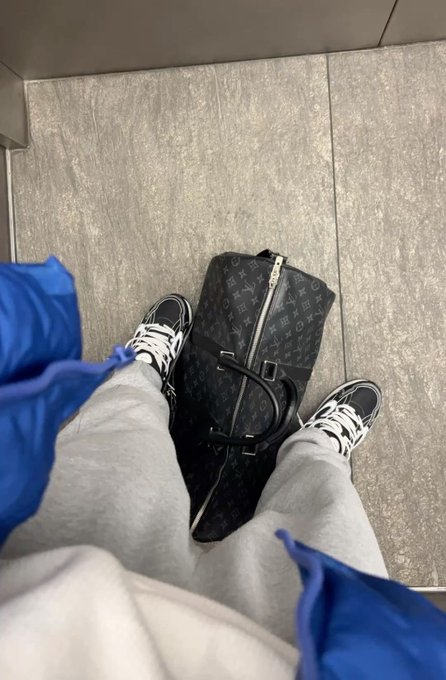 A first-person view of someone wearing grey sweatpants and black and white sneakers, standing over a black Louis Vuitton duffle bag on a tiled floor.