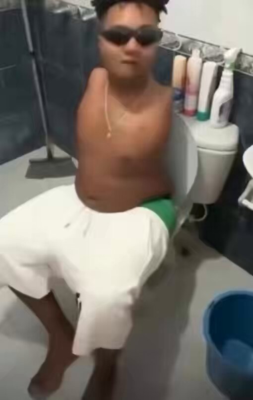 The image depicts a man wearing white swim shorts sitting inside a bathroom, possibly using toilet paper or cleaning his butt with toilet paper rolls. The bathroom features various toiletries such as toothbrushes, bottles, and even a sink. Above the toilet, there is black tile lining the walls of the room. In front of the toilet, there's another blue bucket containing more toilet paper rolls. The image also contains text that reads: "Dude is over here cleaning his ass with his own feet And you can’t even manage a simple click to buy more Solana at $64".