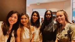 The image shows a group of five women standing together, posing for a photo with smiles on their faces. They are all dressed similarly, wearing white tops that match each other. The background features a brown wall, which complements the color of one woman's dress. In the bottom right corner of the image, there is a caption that reads "solana summer in SF and the highest-value attendee is this @UseCorgi". This caption suggests that the group photo was taken at an event or gathering where the attendees were dressed alike, possibly for a special occasion or celebration.
