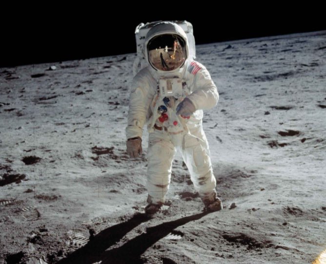 An astronaut in a white spacesuit standing on the gray, cratered surface of the moon with the blackness of space in the background.