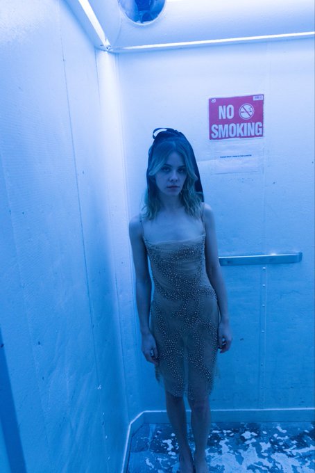 A young woman with blonde hair wearing a sparkly sheer dress and a black bow headband, standing in a blue-lit bathroom with a 'NO SMOKING' sign on the wall behind her.
