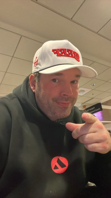 A man wearing a white cap and dark green hoodie with a red logo takes a selfie, pointing his finger toward the camera in an indoor setting.
