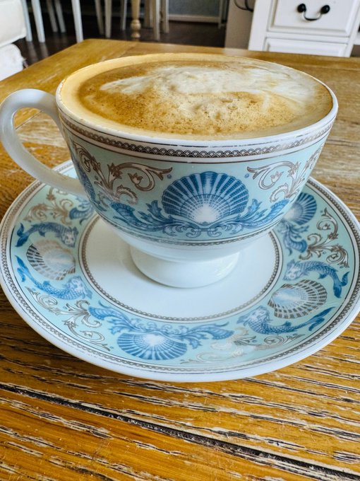An ornate vintage teacup and saucer with intricate blue and white seashell patterns holds a frothy cappuccino dusted with cinnamon, sitting on a rustic wooden table.