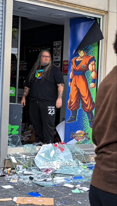 A long-haired, bearded man stands confidently in the shattered doorway of a Dragon Ball Z merchandise shop, surrounded by broken glass and debris with "Nice, France" displayed on a hoodie and "EVERLAST 23" on his sweatpants, projecting an effortlessly unbothered "aura."