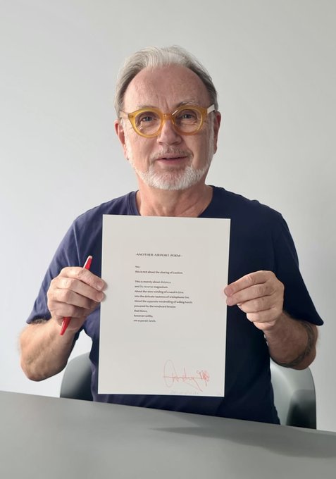 An older man with yellow glasses holds up a poem titled 'ANOTHER SENSES POEM' signed in red marker.
