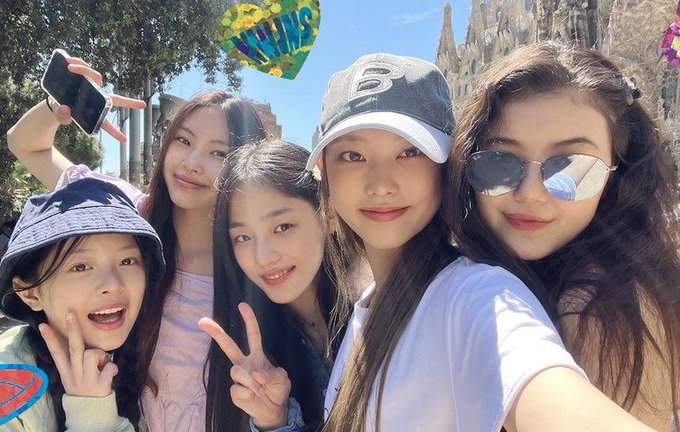 A sunny outdoor group selfie of five young women, one holding a phone and another a fan, posing playfully in front of a wooden structure.