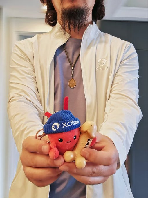 A person wearing a cream-colored XCHL jacket and a wooden pendant necklace holds a red plush toy wearing a blue XCHL beanie and a wooden bone with the text 'XCHL'.