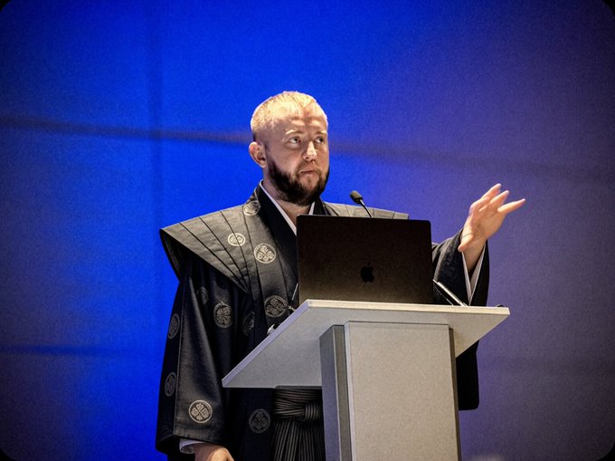A bearded speaker in traditional Japanese attire stands at a podium with a laptop, gesturing mid-presentation. The image illustrates the post's discussion on Chainlink's token release strategy and how governments could serve as trusted nodes to build reputation for future job iterations.