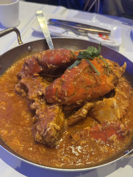 A close-up photo of a large pan filled with chili crab in a rich, thick orange-red sauce, garnished with cilantro, served at a restaurant table.