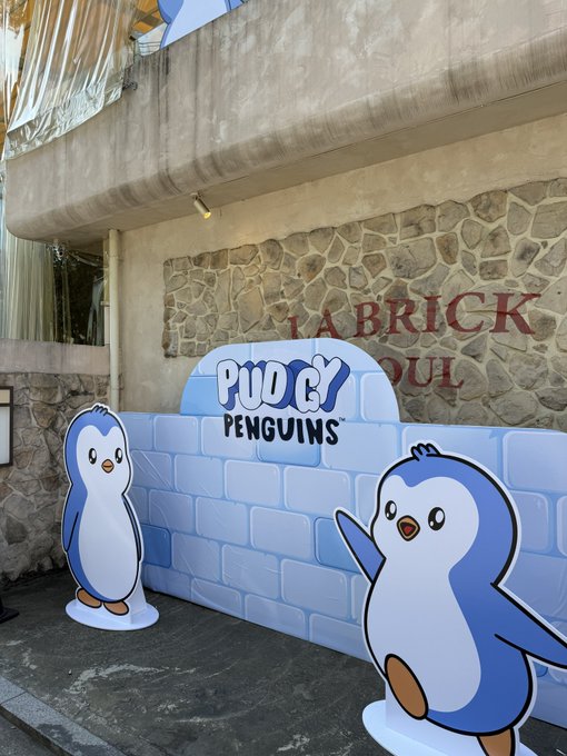 An outdoor photo showing cartoon penguin cutouts and an inflatable 'PUDGY PENGUINS' sign set up in front of a stone wall with 'LA BRICK OUL' text.