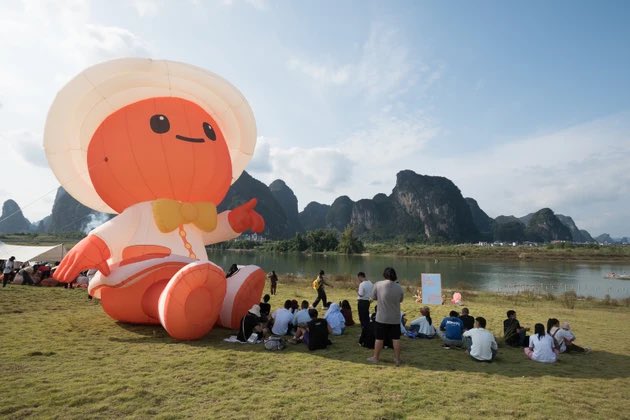 A large orange mascot balloon wearing a white hat and yellow bow tie sits on a grassy field by a river, with people gathered around and dramatic karst mountains in the background.