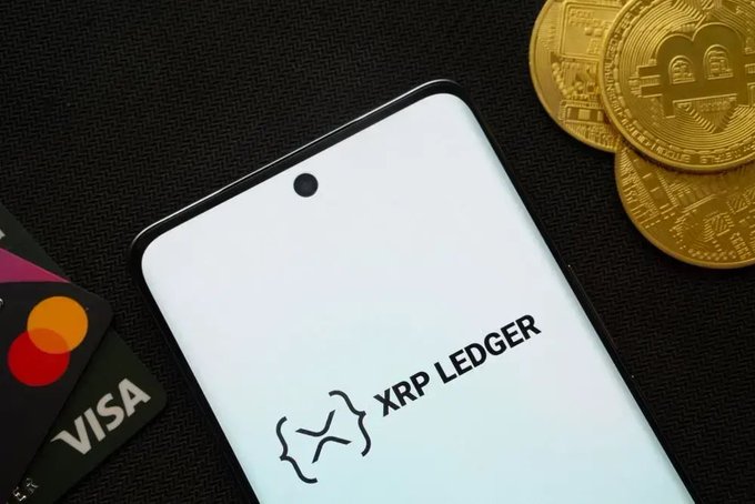 A smartphone displays the "XRP LEDGER" logo, accompanied by gold Bitcoin coins and a Visa/Mastercard credit card on a dark surface. The image visually represents the XRPL hackathon announcement, highlighting the intersection of cryptocurrency development and traditional finance.