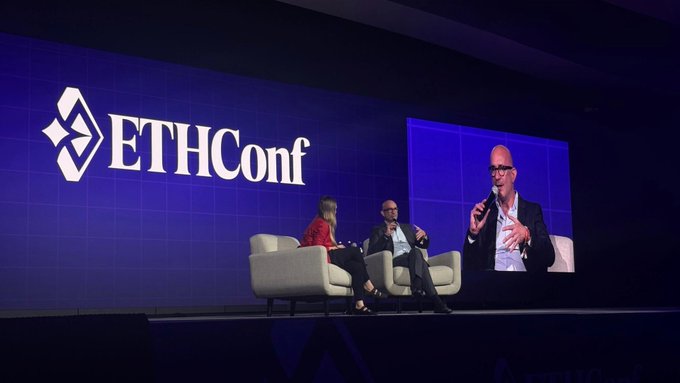 The image shows a stage with two people sitting on chairs, one of whom appears to be speaking into a microphone. The backdrop features a blue wall with the word "ETHConf" written on it. In front of the speakers, there are three chairs arranged in a semi-circle around them. A large screen is also visible behind the speakers, displaying an image or video related to the event.