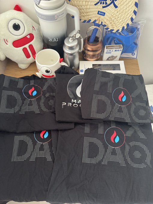 Black merchandise featuring a flame-like logo and the text "DAO PRO" is laid out on a table alongside various other items including a plush toy, thermos, and cup.