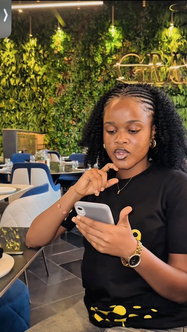 A woman with curly hair wearing a black shirt with a yellow logo, looking at her smartphone while seated in a restaurant with a green plant wall background.