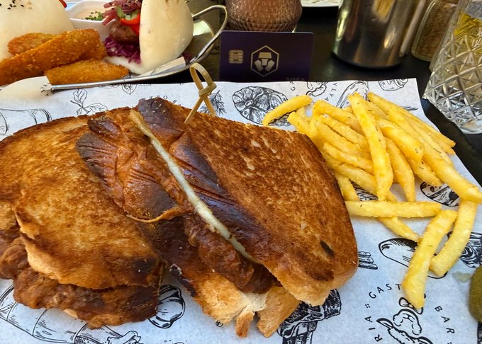 A restaurant meal featuring a toasted sandwich cut in half, french fries, and a starter plate, with a Crypto.com business card visible on the table.