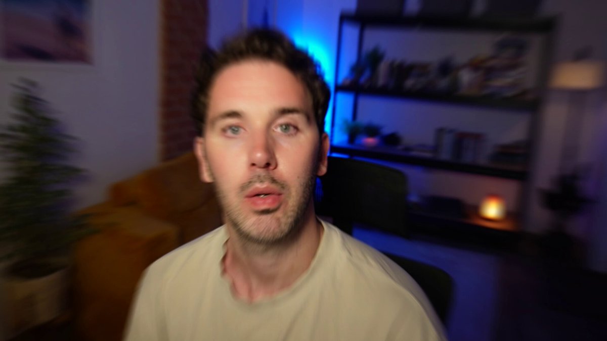 The image shows a man wearing a white shirt sitting in front of a couch with his head tilted back as if he's looking at the camera or posing for a photo. The background features a bookshelf filled with various items, including several books on different shelves and a potted plant placed near one of them. There is also an image of a candle on the bookshelf that appears to be lit.