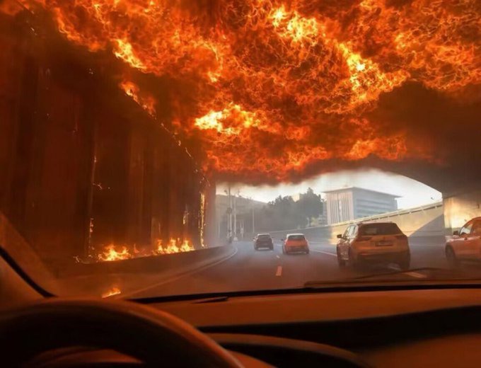 The image shows a car driving down a road with a large plume of smoke coming from behind, possibly due to an accident or fire. The sky above appears to be ablaze, creating a dramatic backdrop for the scene. In the foreground, there is a car's windshield and a person standing nearby. A post on social media reads: "let me out of my longs i dont even want a profit ill do anything".