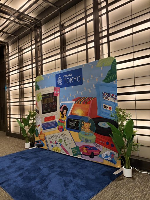 An 'EthGlobal TOKYO' event sign with colorful illustrations of computers, cars, and robots, placed on a blue rug in front of wooden slatted walls.