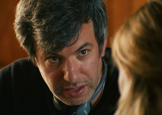 A close-up of a man with dark hair wearing headphones around his neck, looking intently toward a blonde-haired person in the foreground.