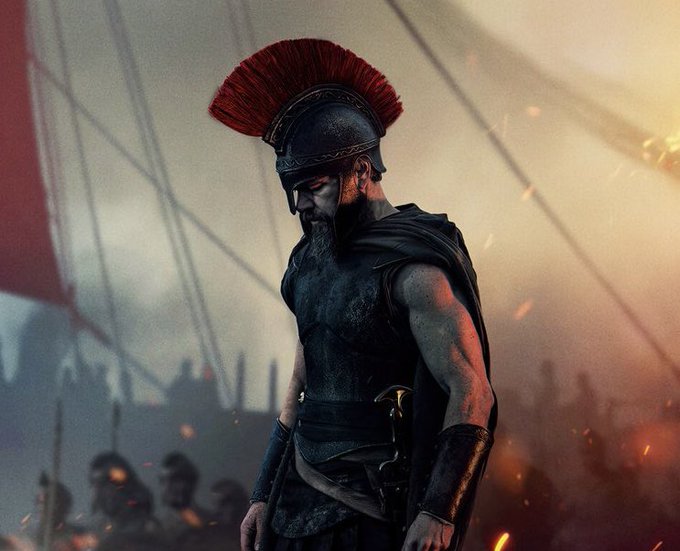 A cinematic image of a bearded warrior wearing a red-plumed Roman or Spartan helmet and dark armor, standing amid a smoky battlefield with ships and embers in the background.