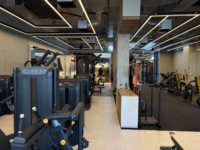 An empty, modern industrial-style gym featuring black weight machines, dumbbells, linear LED ceiling lights, and a central wooden bench.