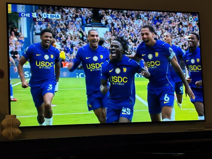 Chelsea soccer players in blue "USDC by Circle" sponsored jerseys celebrate a goal during a Premier League match against Brighton (CHE vs BHA, 04:53), displayed on a TV screen.