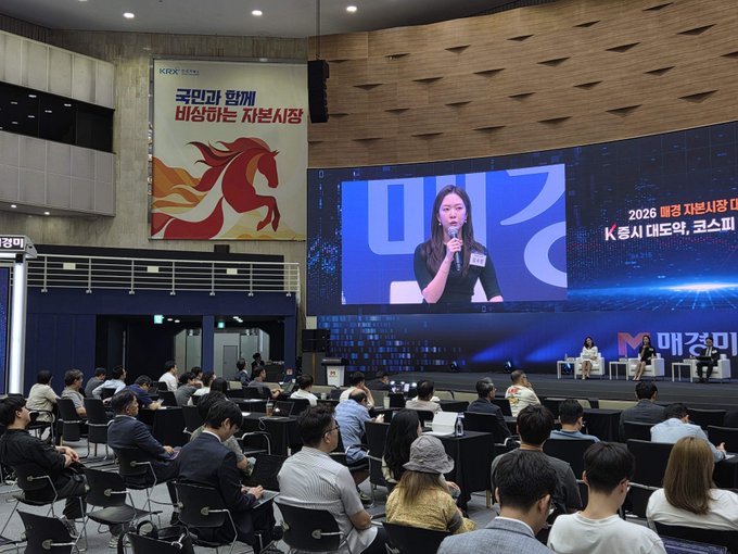 The image shows a large group of people sitting in chairs, watching a presentation or event taking place on a big screen TV. The audience appears engaged and attentive as they listen to the speaker. The room has a modern design with blue walls that create an inviting atmosphere for the gathering. The attendees are seated at various positions within the room, some closer to the front while others are further back. Overall, it is a scene of people coming together to learn or enjoy an event in a comfortable setting.
