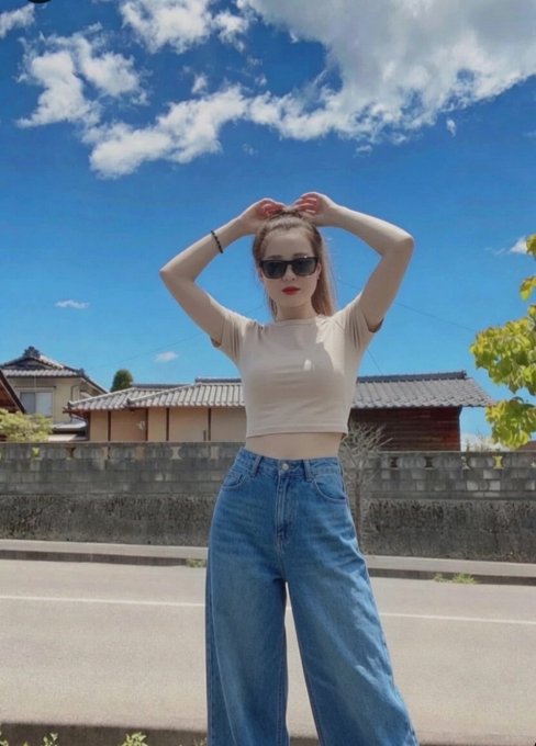 A photograph of a young woman wearing a beige crop top, blue jeans, and sunglasses, posing with her hands near her head on a sunny day, with traditional Japanese houses, a stone wall, and a bright blue sky in the background.