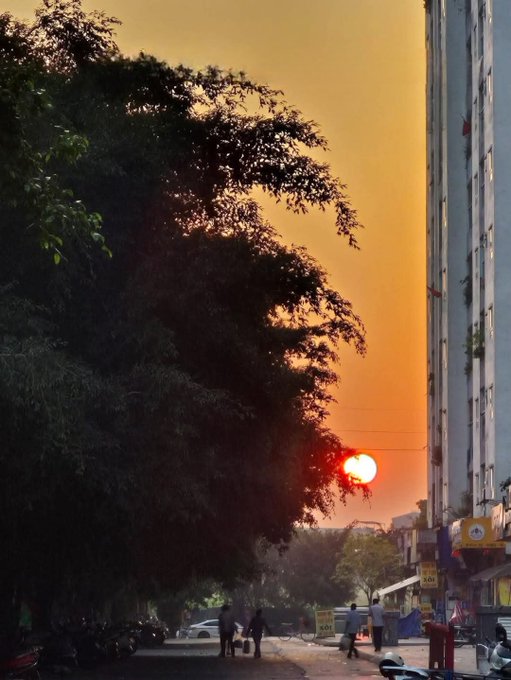 A photograph of a vibrant orange sunset over a city street, with silhouettes of large trees on the left, a tall apartment building on the right, and people walking below.
