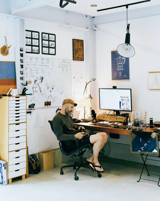 A bearded man in a cap and shorts sits at a cluttered desk in a bright studio space, working on a computer displaying a 'Mirokoshi' webpage, with artwork and sketches pinned to the walls.