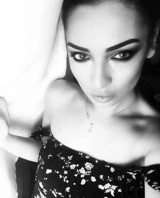 A striking black and white close-up selfie of a woman with dramatic eye makeup, wearing a floral off-the-shoulder top and a delicate necklace.