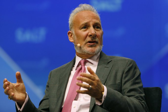 An older man with gray hair and beard, wearing a gray suit and pink tie with a headset microphone, gestures with both hands while speaking against a blue background.