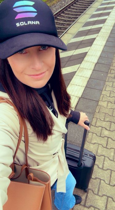The image features a woman wearing a black baseball cap with a blue logo, standing next to a train station platform. She has her handbag over her shoulder and appears to be waiting for transportation or possibly just passing by. The caption of the image reads "GM see you at solana summit germany?". This suggests that the photo might have been taken during an event related to Solana, such as a conference or gathering, where the woman is either attending or has attended.