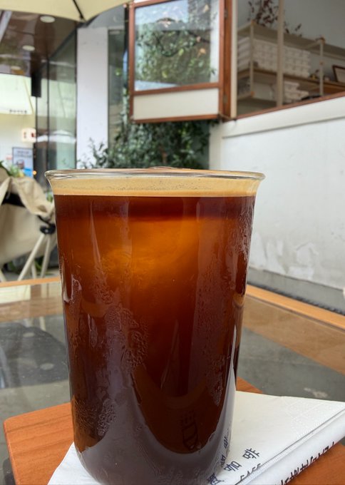 A glass of iced coffee or cold brew beverage on a wooden table at what appears to be a café, with a napkin bearing Chinese characters visible beside it.