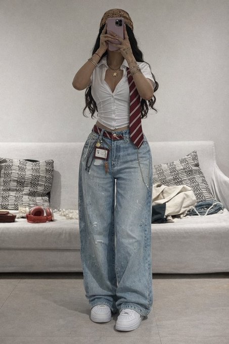 The image features a woman standing in front of a couch, wearing blue jeans with a white shirt and a red tie. She has her arm up in the air while taking a selfie using her phone. The background includes a couch and some pillows, creating a casual atmosphere for the photo.