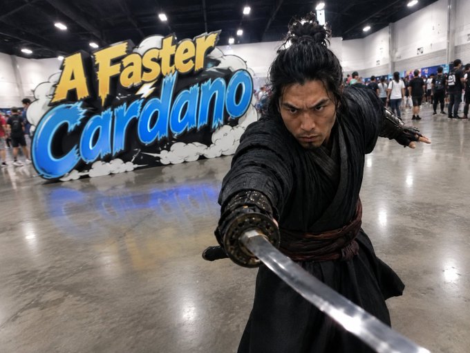 A man dressed as a samurai wields a katana in a dynamic pose in front of a large 'A Faster Cardano' comic-style sign at a convention.