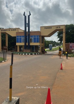 The entrance gate of the "University of Ilorin," featuring a yellow archway, a security post, a paved road with black and white curbs, and a yellow bollard with the text "Swipe up for filters" overlaid.