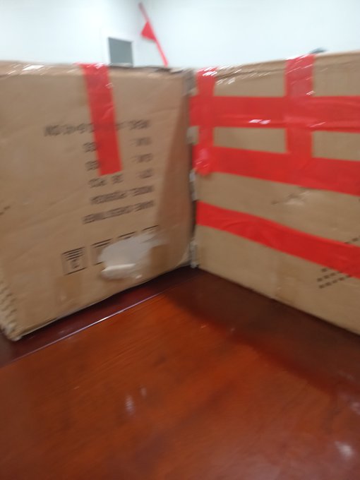 Two large cardboard boxes sealed with red tape sit on a wooden floor, with printed product details visible on the side.