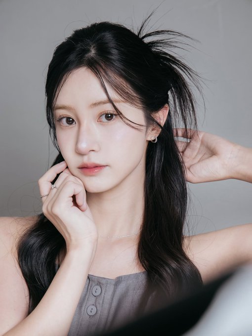 A close-up portrait of a woman with long dark hair, wearing a gray buttoned top, posing with her hands near her face against a plain background.
