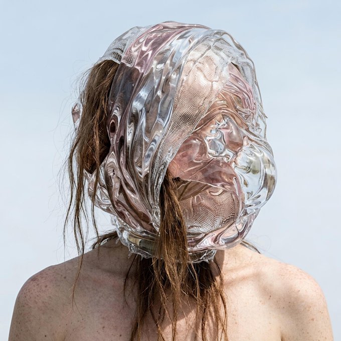 A back view of a person wearing an elaborate, sculptural clear vinyl hood or headpiece with mesh fabric, with wet-looking brown hair emerging from underneath against a pale blue background.