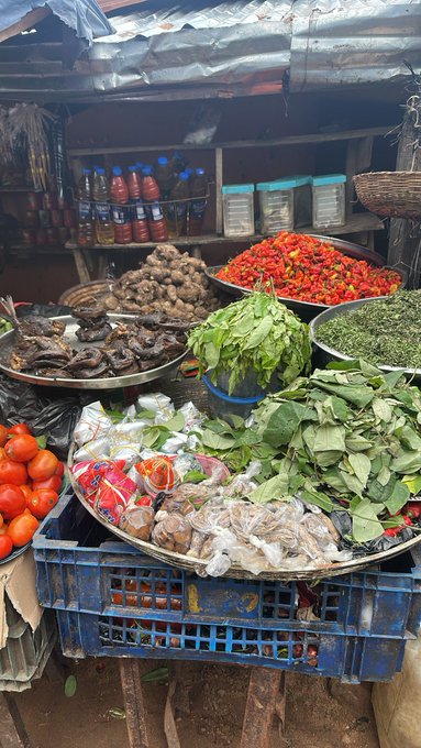 A vibrant outdoor market stall displays fresh produce including red tomatoes, green leafy vegetables, ginger root, and dried fish, alongside jars of spices and bottles in the background. The image connects to the post by showcasing a variety of ingredients that could inspire a home-cooked meal during the user's relaxing Saturday, paired with their plan to binge-watch *Black Torch*.