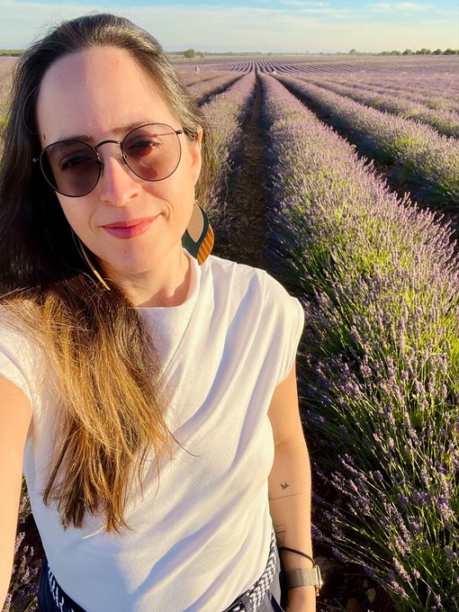 A woman wearing sunglasses and a white t-shirt takes a selfie in front of a vibrant field of blooming lavender at sunset.