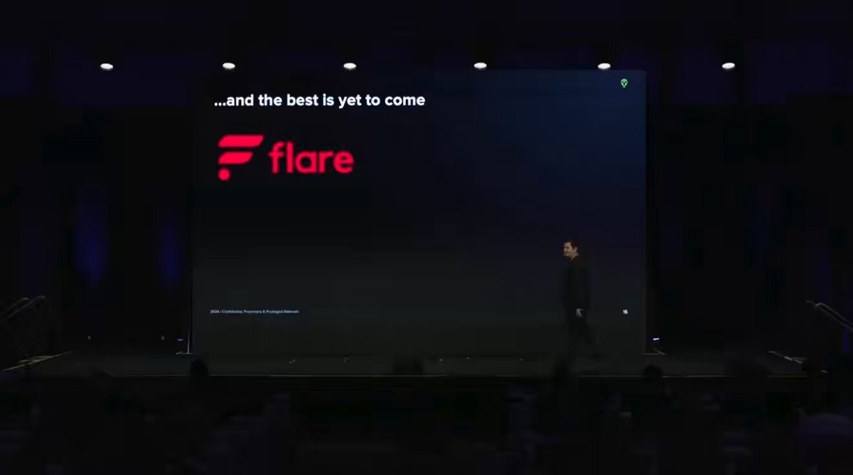 The image shows an event stage with a large screen displaying the words "And the best is to come" along with a man standing behind the screen, possibly giving a speech or presentation. The background of the image features a dark room with multiple chairs arranged in rows and a few people sitting among them.
