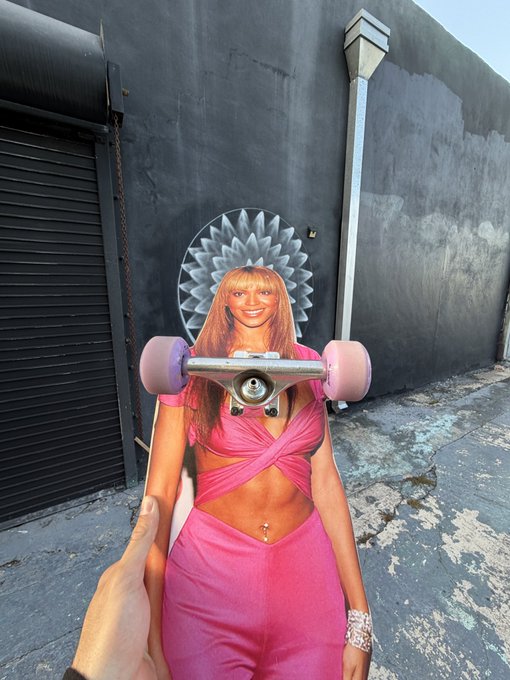 A cardboard cutout of a smiling woman in a pink outfit being held up outdoors on a city sidewalk, with a skateboard appearing to be balanced across her cutout neck.