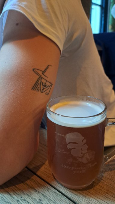 A close-up of a person's leg bearing a small line-art tattoo of a face wearing a hat, next to a glass of amber beer on a wooden table.
