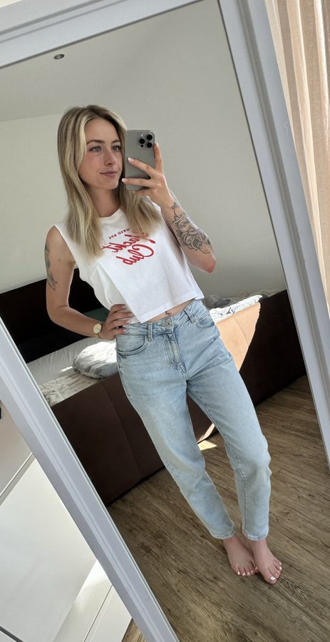 A mirror selfie of a blonde woman wearing a cropped white t-shirt and light blue jeans, showing tattoos on her left arm, taken in a bedroom with a bed visible in the reflection.