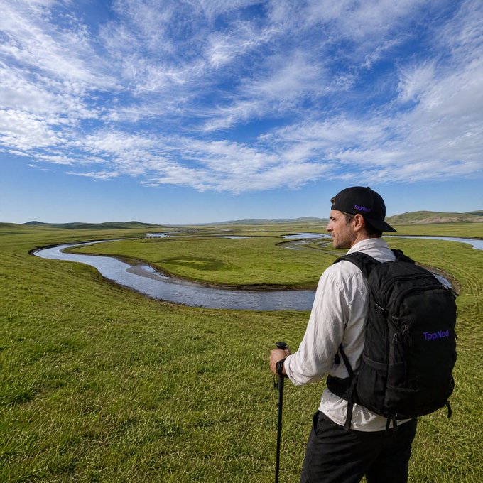 A man wearing a black 'TopNod' backpack and matching cap gazes out over a winding river flowing through a vast green grassy plain under a blue, cloudy sky.